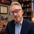 A person with short gray hair and a beard, wearing tortoiseshell glasses and a navy blazer over a light blue dress shirt. They are seated in an office decorated with Syracuse University memorabilia
