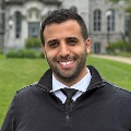 A smiling individual standing in front of a historic stone building.
