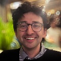 Portrait of a smiling person with glasses, wearing a black and white checked shirt under a sweater.