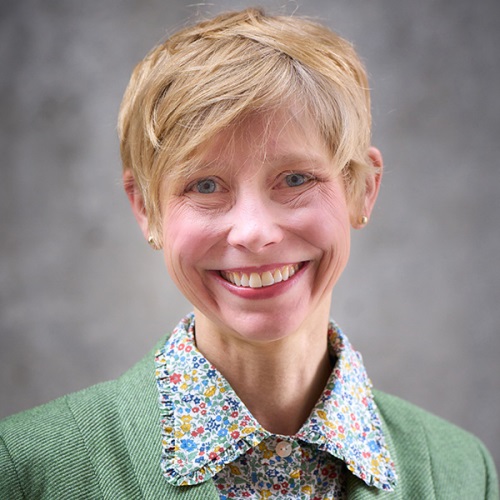 A portrait of a person smiling, wearing a green sweater and a floral collar shirt, set against a plain gray background.