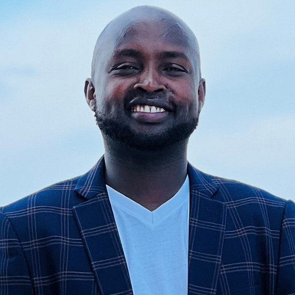 A person smiling outdoors against a light blue sky. They are wearing a navy plaid blazer over a white v-neck shirt.