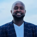 A person smiling outdoors against a light blue sky. They are wearing a navy plaid blazer over a white v-neck shirt.