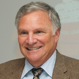 Smiling person with gray hair, tweed blazer, and patterned tie.
