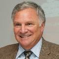 Smiling person with gray hair, tweed blazer, and patterned tie.