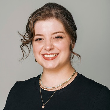 Professional portrait of a smiling person with brown hair wearing a black top and several necklaces.