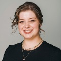 Professional portrait of a smiling person with brown hair wearing a black top and several necklaces.