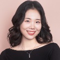 Person with wavy hair, black off-shoulder top, and a pearl necklace against a pink backdrop.