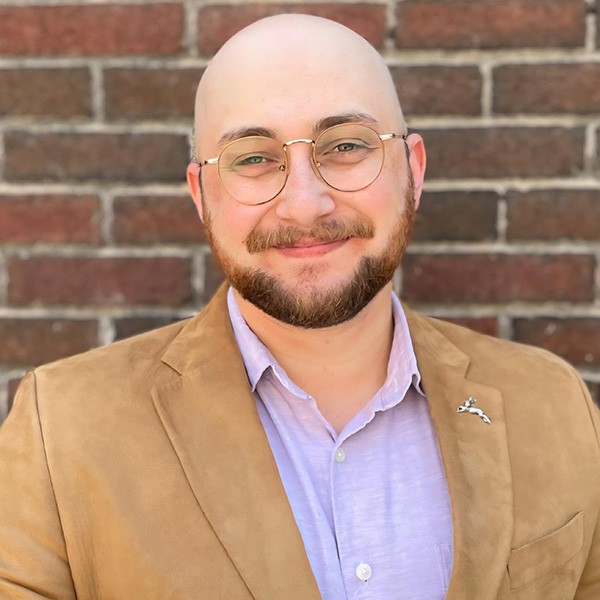 Portrait of a smiling individual in a brown blazer and light blue shirt, standing against a brick wall background.