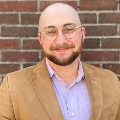 Portrait of a smiling individual in a brown blazer and light blue shirt, standing against a brick wall background.