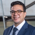 Photo of a smiling person with dark hair and glasses wearing a blue suit and tie