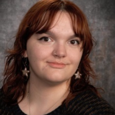 A person with reddish brown hair and brown eyes wearing star earrings and a black top.