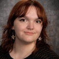 A person with reddish brown hair and brown eyes wearing star earrings and a black top.