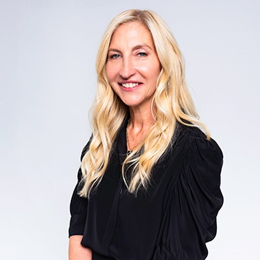 A person with long blonde hair, wearing a black top, smiling against a light gray background.