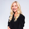 A person with long blonde hair, wearing a black top, smiling against a light gray background.