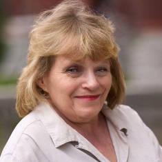 Portrait of a smiling person with blonde hair, wearing a light beige shirt, set against a blurred background.