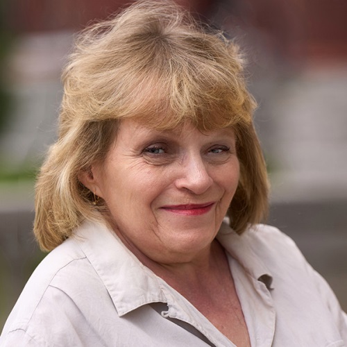 Portrait of a smiling person with blonde hair, wearing a light beige shirt, set against a blurred background.