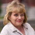 Portrait of a smiling person with blonde hair, wearing a light beige shirt, set against a blurred background.