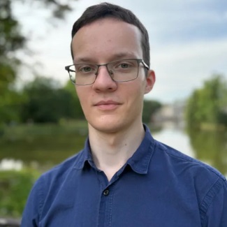 Close-up portrait of a person wearing glasses and a blue shirt, standing outdoors with a blurred river and trees in the background.