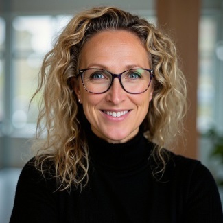 A person with blonde, curly hair smiling and wearing a black turtleneck top and glasses.