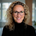 A person with blonde, curly hair smiling and wearing a black turtleneck top and glasses.