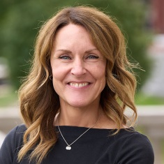 Portrait of a smiling person with long, wavy brown hair, wearing a black top and a small pendant necklace, with a blurred green background.