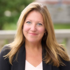Portrait of a smiling individual with long blonde hair, wearing a black jacket and white top, set against a softly blurred outdoor background.