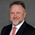 A person with short gray-blond hair and a neatly trimmed beard smiles at the camera, wearing a dark suit jacket and a red tie against a gray studio background.