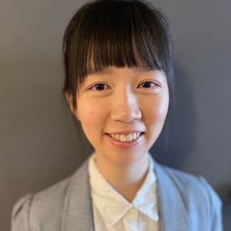 Portrait of a smiling person wearing a grey blazer and white shirt, in soft indoor lighting.