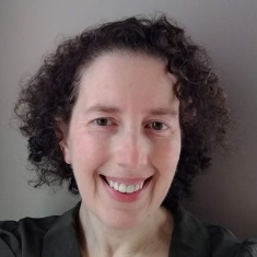 A person with short, curly dark hair smiles warmly at the camera against a neutral gray background. They is wearing a dark olive green top.