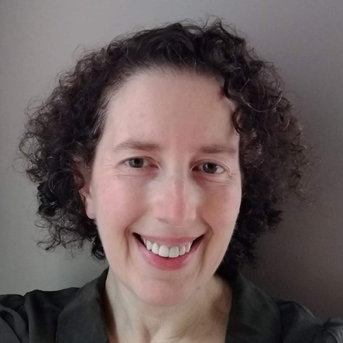 A person with short, curly dark hair smiles warmly at the camera against a neutral gray background. They is wearing a dark olive green top.