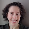 A person with short, curly dark hair smiles warmly at the camera against a neutral gray background. They is wearing a dark olive green top.