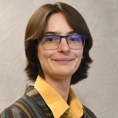 Portrait of a smiling individual wearing glasses, a plaid blazer, and a yellow shirt against a light background.