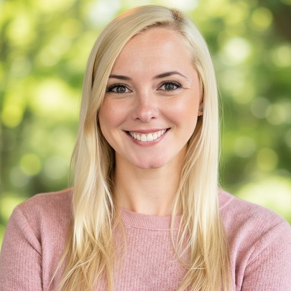 Portrait of a smiling person with blonde hair, wearing a pink sweater. The background is softly blurred with green tones.