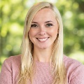 Portrait of a smiling person with blonde hair, wearing a pink sweater. The background is softly blurred with green tones.