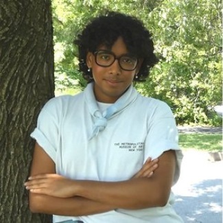 Photo of a person wearing glasses with their arms crossed and leaning against a tree.