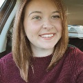 Close-up portrait of a smiling individual with medium-length brown hair, wearing a burgundy sweater, sitting in a car.