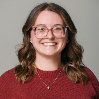 A smiling person with wavy hair, glasses, and a maroon sweater.