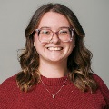 A smiling person with wavy hair, glasses, and a maroon sweater.