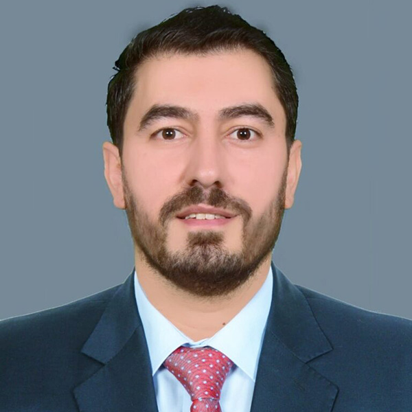 Professional headshot of a person wearing a dark blue suit, white shirt, and red patterned tie, against a grey background.