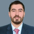 Professional headshot of a person wearing a dark blue suit, white shirt, and red patterned tie, against a grey background.