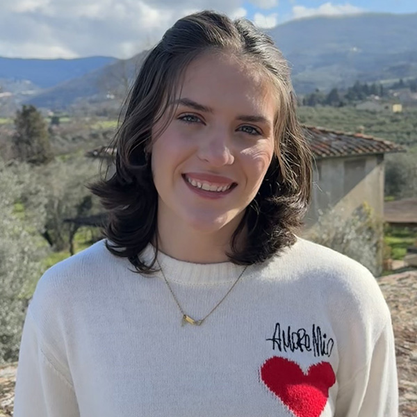 Person in a white sweater with a heart design, smiling with a scenic landscape of hills and foliage in the background.