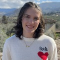 Person in a white sweater with a heart design, smiling with a scenic landscape of hills and foliage in the background.
