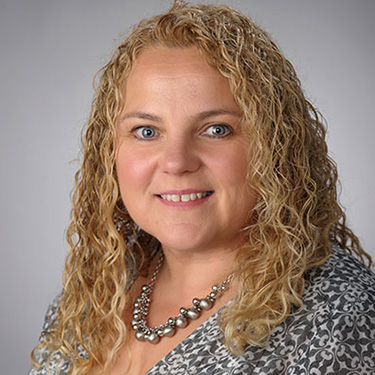 Portrait of an individual with curly blonde hair, wearing a patterned top and a silver necklace, smiling at the camera.