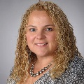 Portrait of an individual with curly blonde hair, wearing a patterned top and a silver necklace, smiling at the camera.