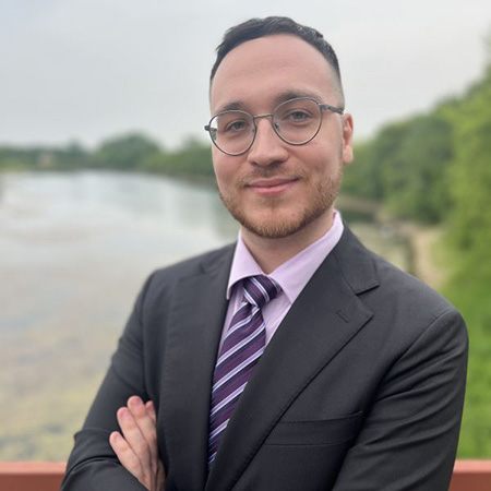 Professional portrait of a person wearing glasses, a suit, and a striped tie, standing by a lakeside with trees in the background.