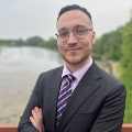 Professional portrait of a person wearing glasses, a suit, and a striped tie, standing by a lakeside with trees in the background.