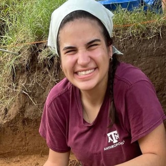 A person with a joyful expression wearing a maroon Texas A&M University Anthropology t-shirt and a white headscarf, kneeling by a dirt excavation site.