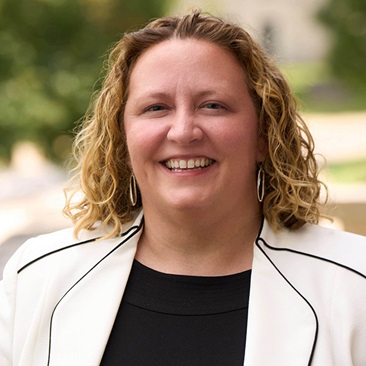 Portrait of a smiling individual with curly hair, wearing a black outfit with a white-trimmed blazer.