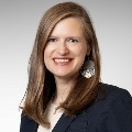 Portrait of an individual smiling, wearing a navy blazer and decorative earrings.