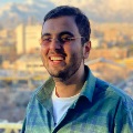 Portrait of a smiling person wearing eyeglasses and a plaid shirt, with a cityscape in the background at sunset.